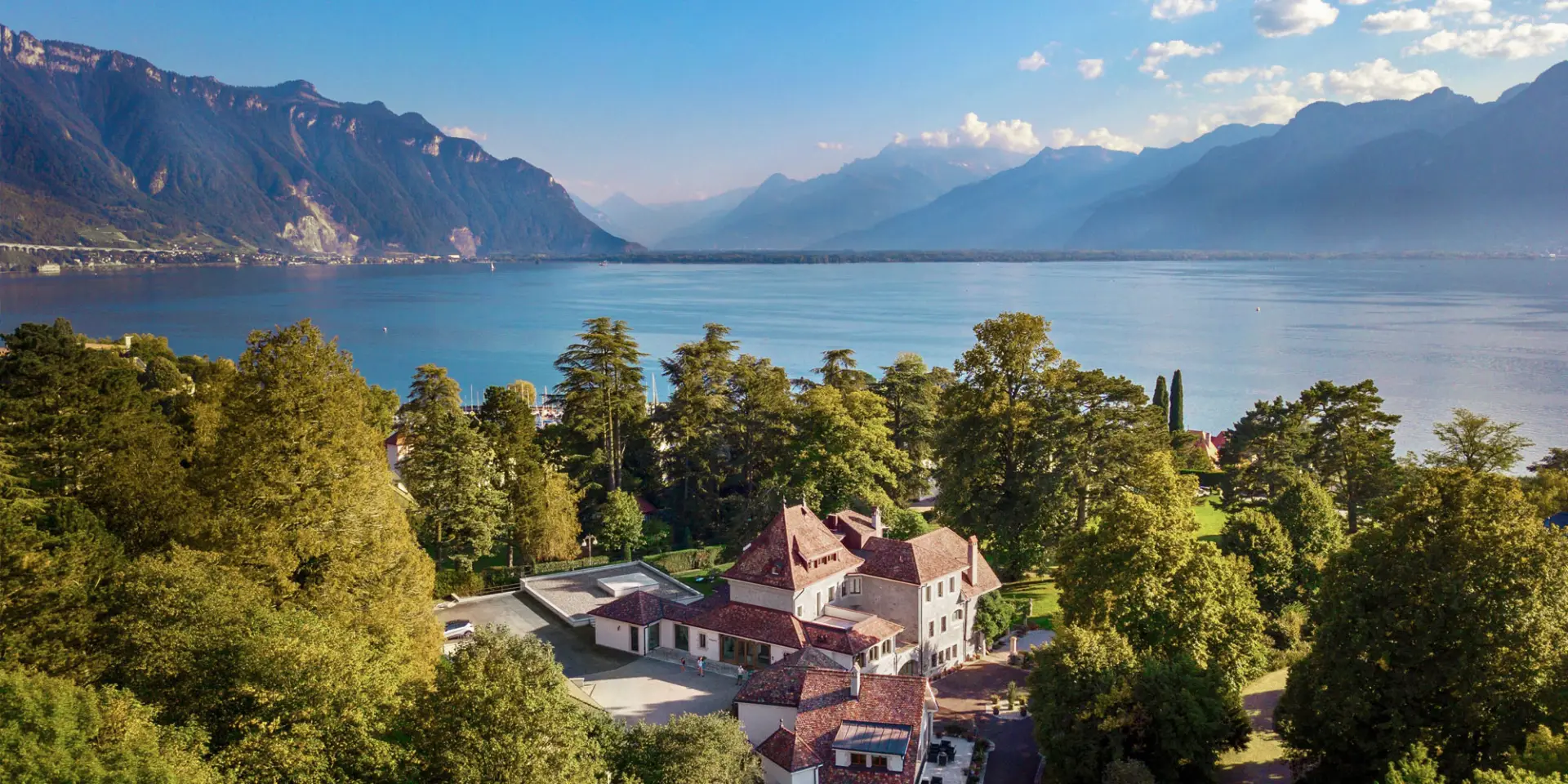 Aerial view of a GuestLee luxury rental in Switzerland, nestled by trees, lake, and mountains under a blue sky. | GuestLee luxury rental Switzerland