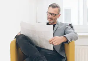 LEE_NewPaper GuestLee luxury rental: A man in glasses and a gray blazer reads in a sunlit armchair, enjoying Swiss comfort. | GuestLee luxury rental Switzerland