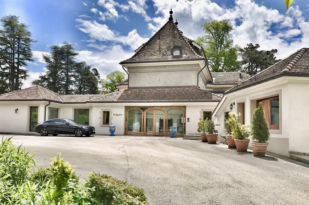 Elegant white Geneva villa with arched entrance, luxury rental by GuestLee, potted plants, black car, Swiss sky backdrop. | GuestLee luxury rental Switzerland