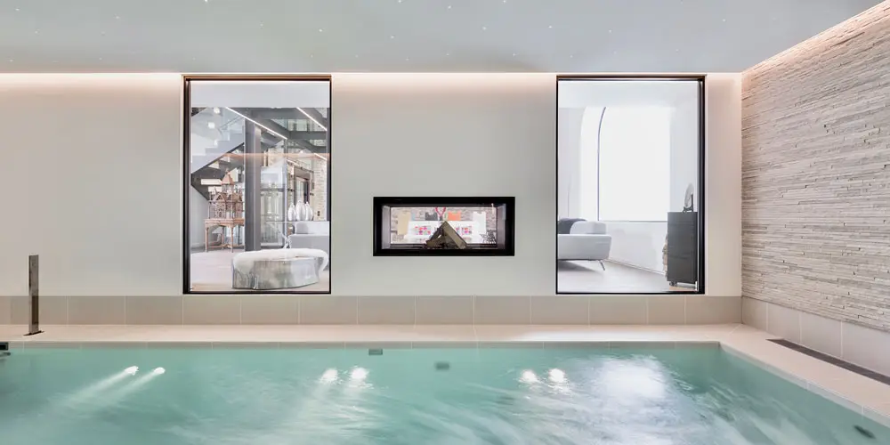LEE_Owner_2 Luxurious indoor pool with blue water, chic tiles, fireplace, and large windows in a Geneva GuestLee villa rental. | GuestLee luxury rental Switzerland