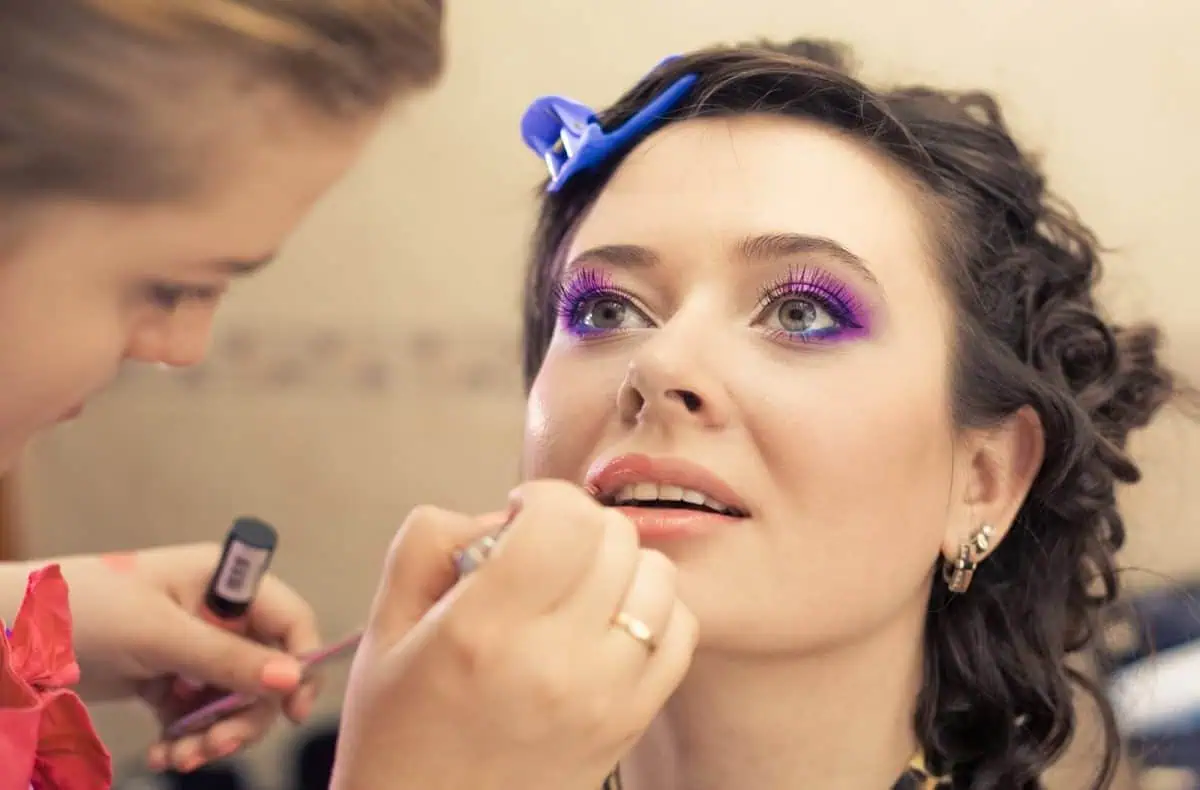 A woman with bright purple eye makeup is having lipstick applied by a hired makeup artist.