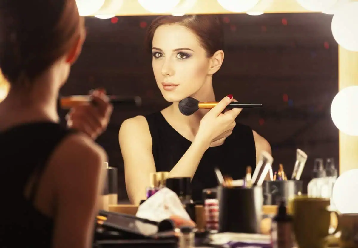 In her cozy dressing room, a woman prepares her GO Bag for a modeling gig, thoughtfully applying makeup with a brush in front of the mirror, as an array of cosmetics surrounds her.