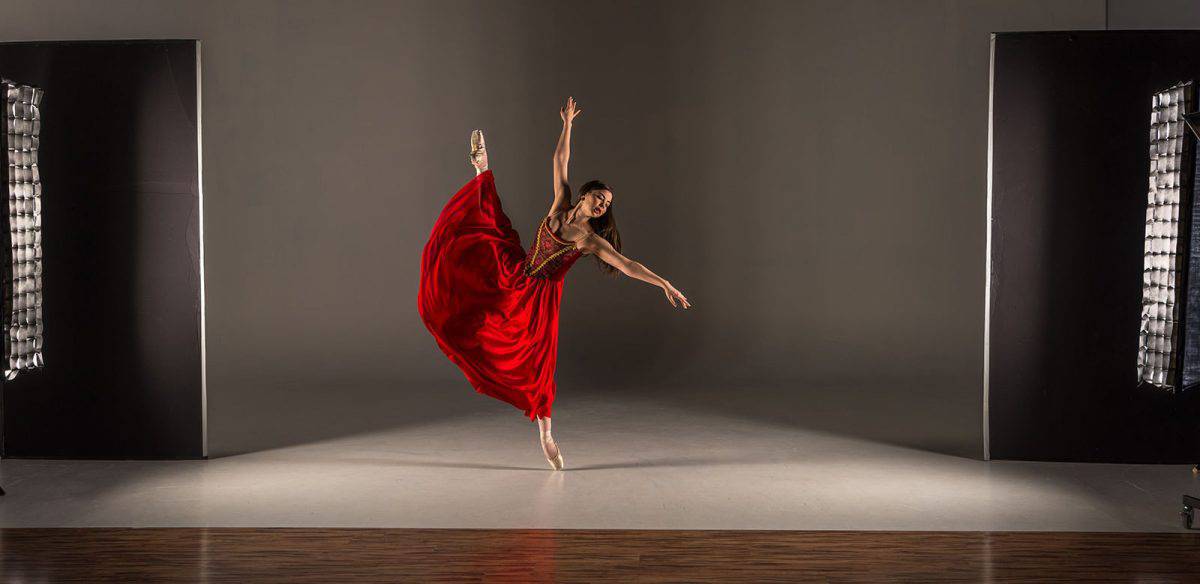 A ballet dancer in a red dress, leaping gracefully on stage between two large lights, creates a moment worthy of any modeling portfolio.