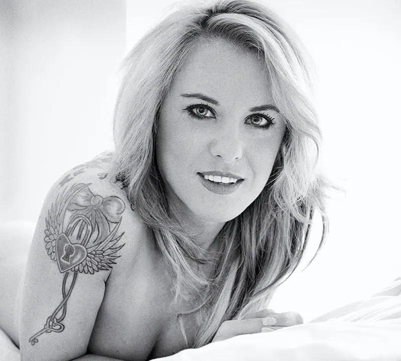 Black and white image of a person with long hair, modeling with tattoos, lying on a bed and gazing confidently at the camera.