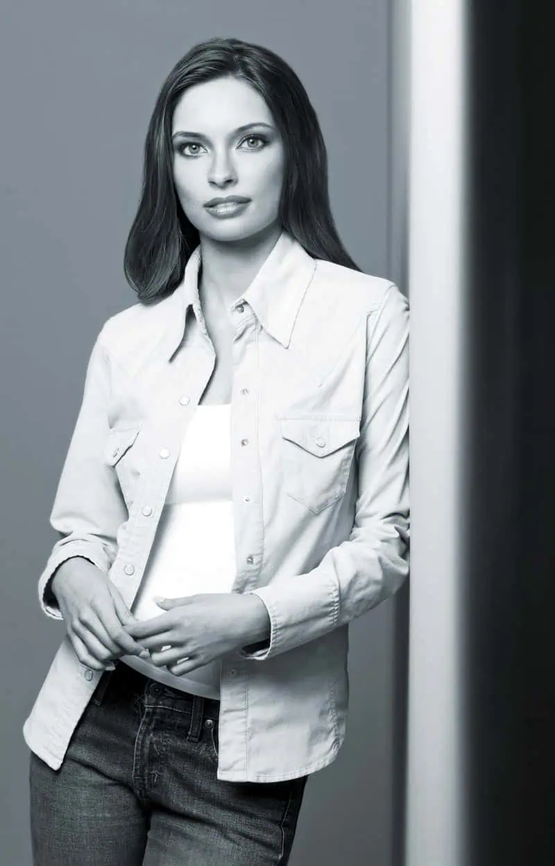 A woman in a denim shirt and jeans stands against a wall, capturing a casual portrait with her neutral expression as she gazes forward.