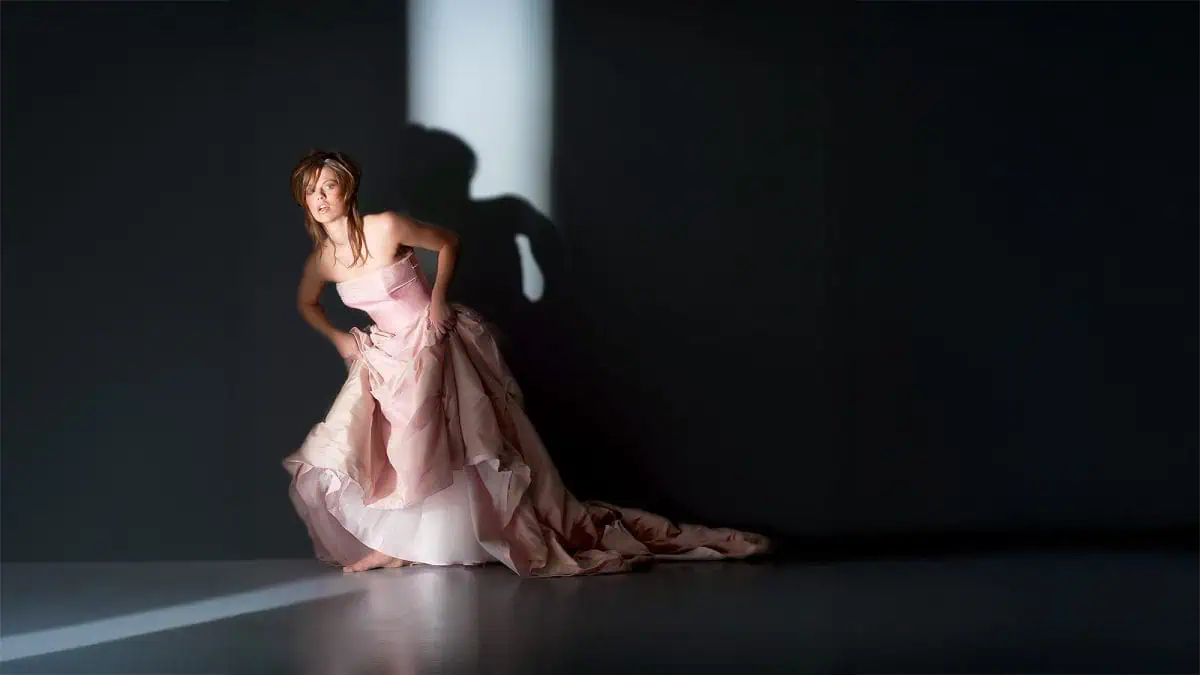 A woman in a pink gown stands against a dark wall, partially lit by a vertical beam of light created with DIY gobos, casting a striking shadow behind her.