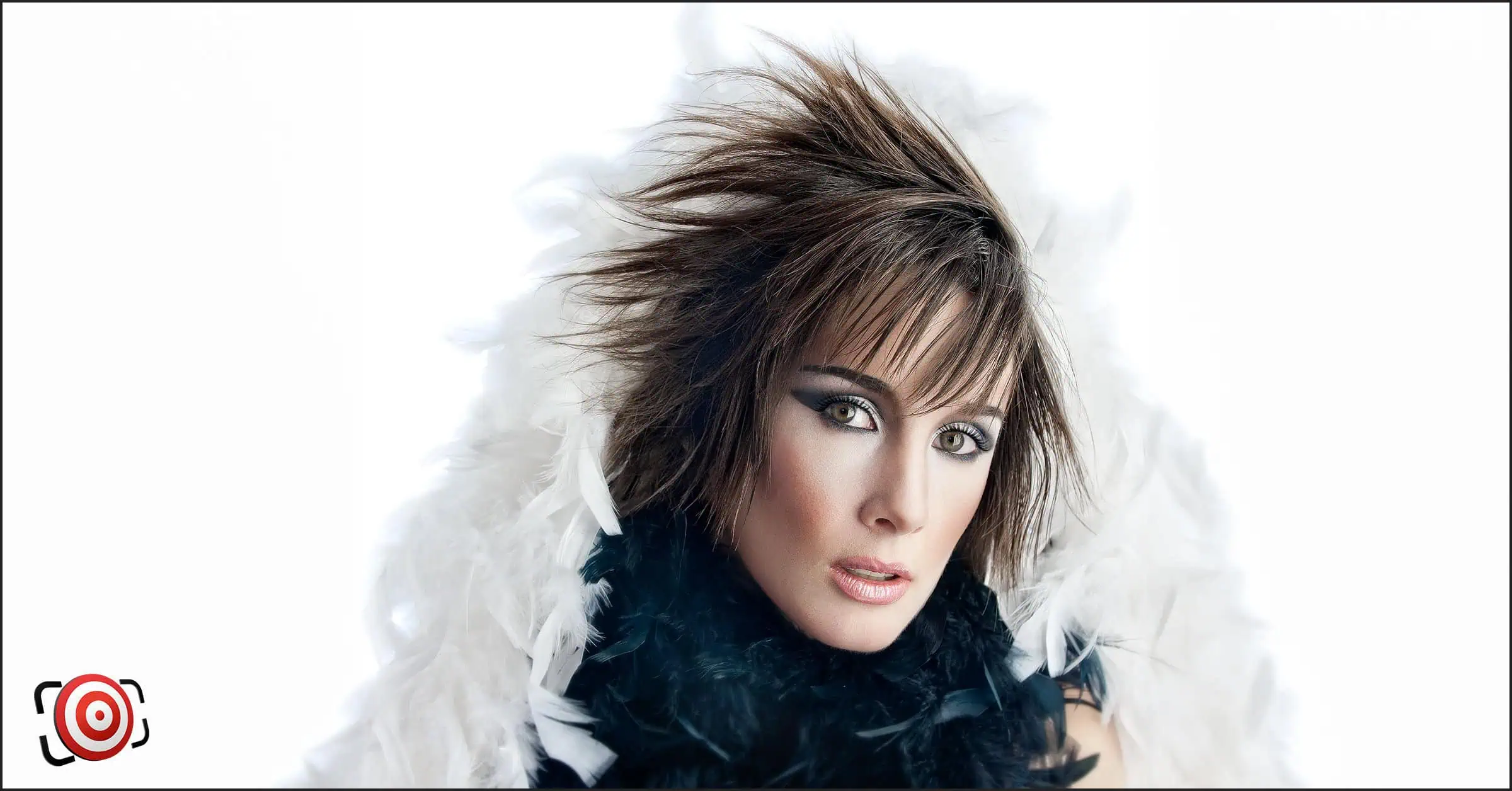 A person wearing dramatic makeup and a feathered outfit with spiked hair poses against a white background, enhanced by a striking lighting arrangement.