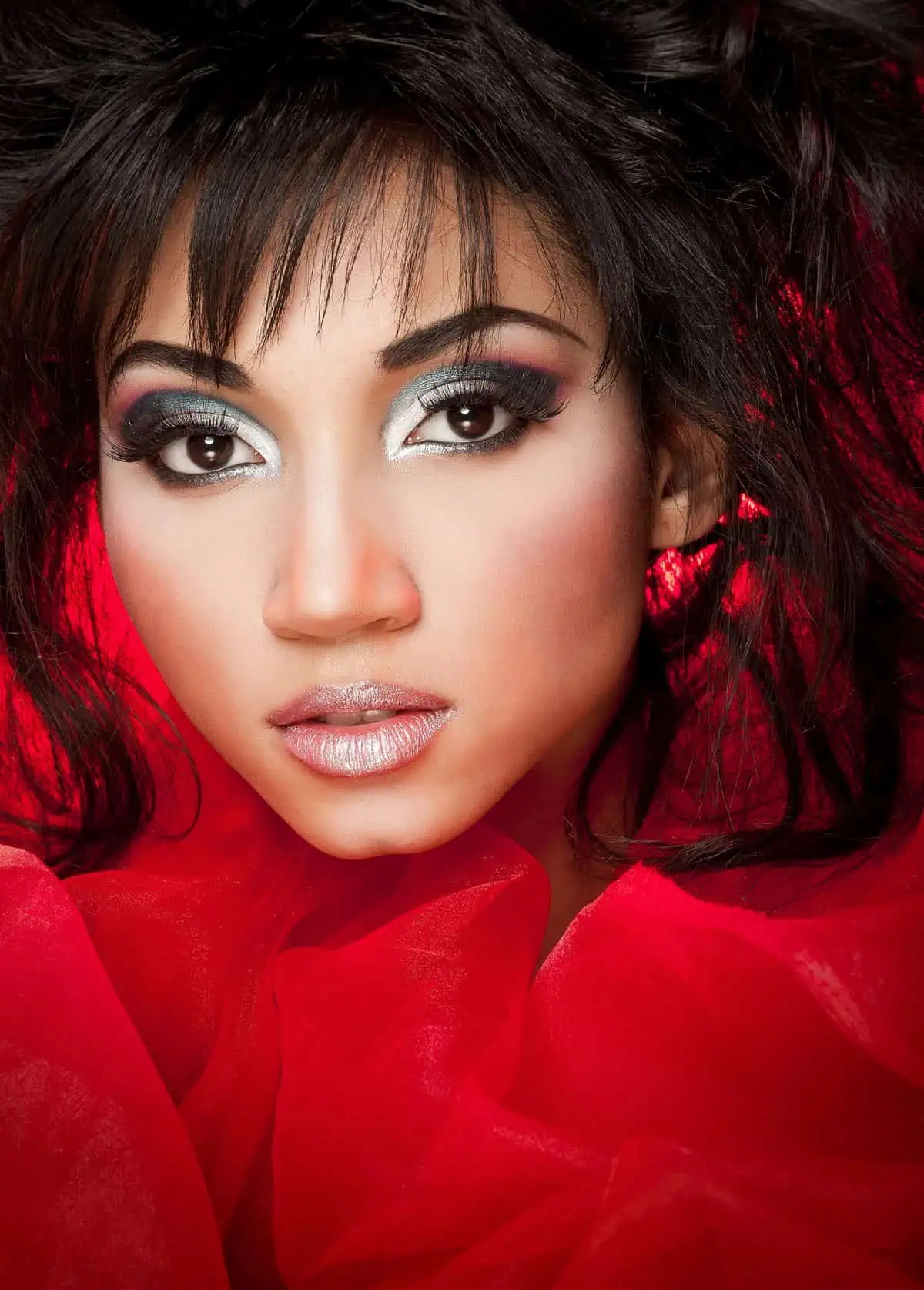 A woman, framed by a red fabric background, gazes intensely at the camera. Her dramatic makeup, highlighted under the soft glow of a beauty dish, adds an enchanting allure to the scene.