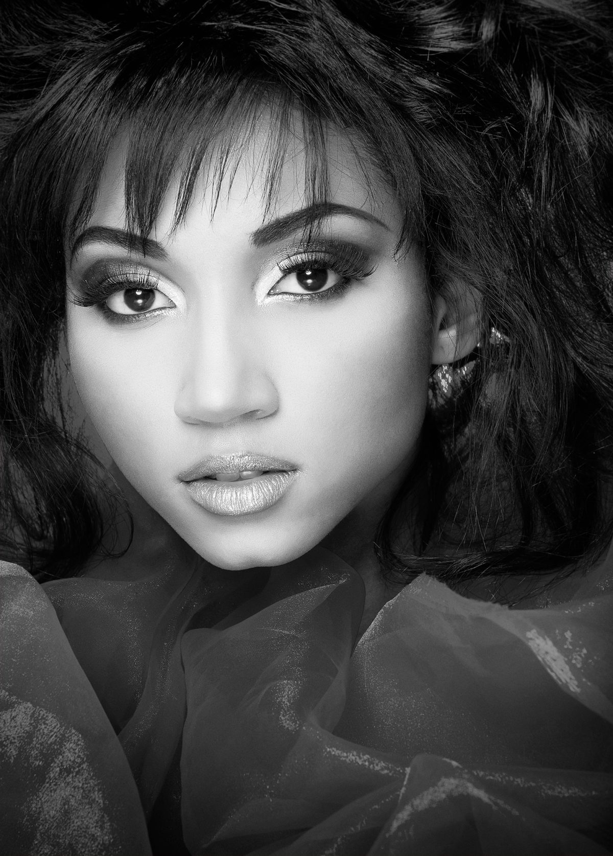 Black and white portrait of a person with dramatic eye makeup and textured hair, illuminated by a beauty dish, looking directly at the camera, framed by soft fabric folds.