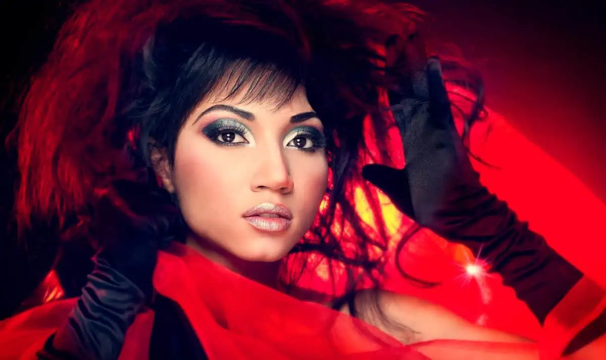 Woman with dramatic makeup and long black gloves poses under a beauty dish in front of a red background, with her hands near her face.