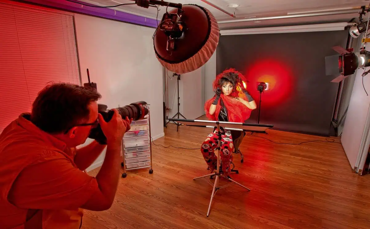 The photographer skillfully captures a model in a studio, enhanced by the dramatic red lighting and a beauty dish that adds depth to the scene, with various equipment subtly visible in the background.