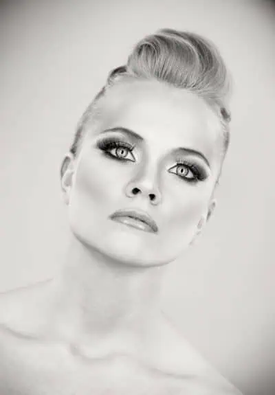 Black and white portrait of a person with light eyes, dramatic makeup, and styled hair, looking slightly upward with a neutral expression against a plain background—an inspiring way to Improve Your Photography skills.