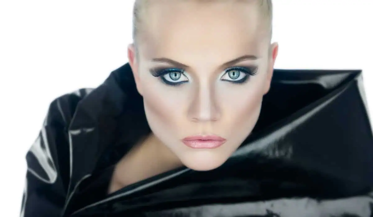 Close-up of a person with blue eyes, dramatic makeup, and light hair, wearing a shiny black garment against a white background—perfect inspiration to improve your photography.