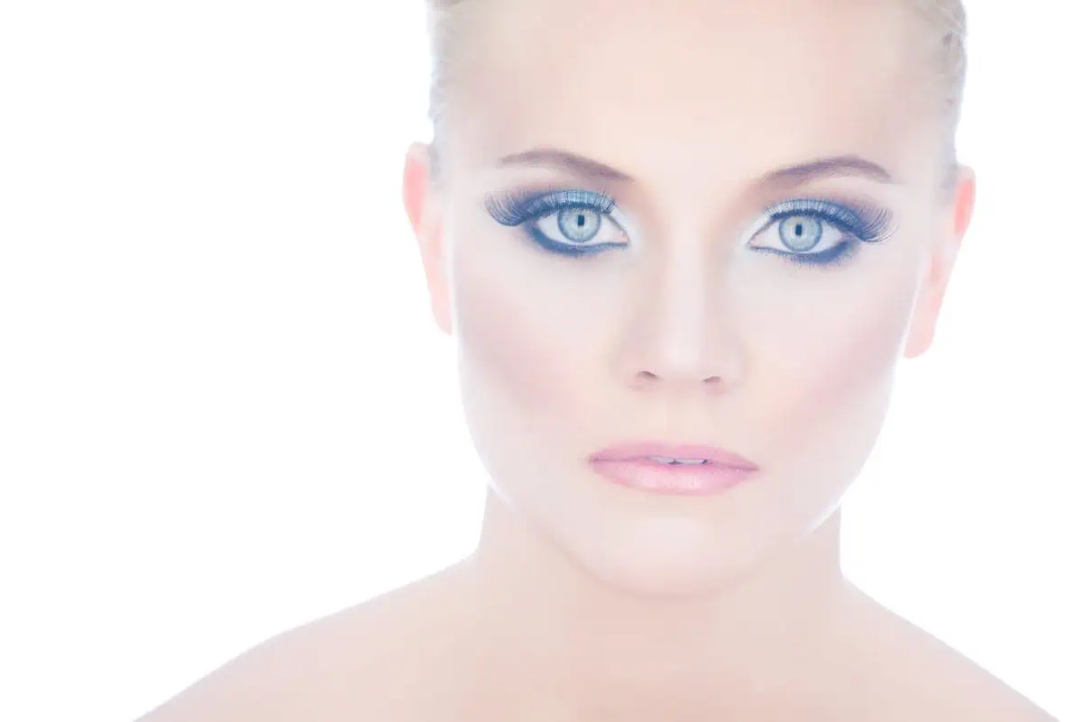 Close-up of a woman with pale skin, blue eyes, heavy eye makeup, and nude lipstick against a bright white background—perfect inspiration to improve your photography with striking portrait techniques.