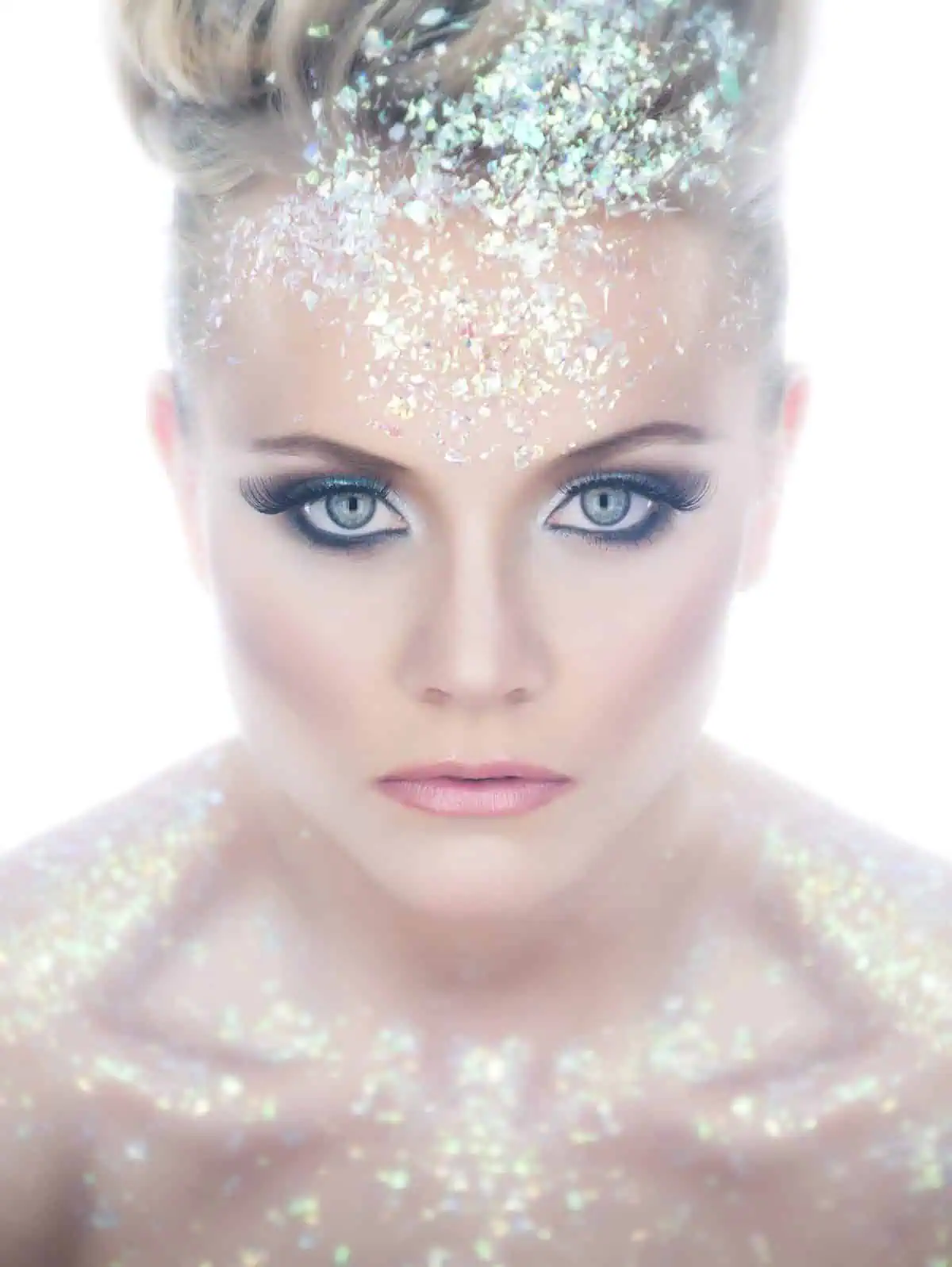 Woman with intense eye makeup stares forward, her face and shoulders covered in iridescent glitter flakes against a bright white background—an inspiring shot to help you improve your photography techniques.