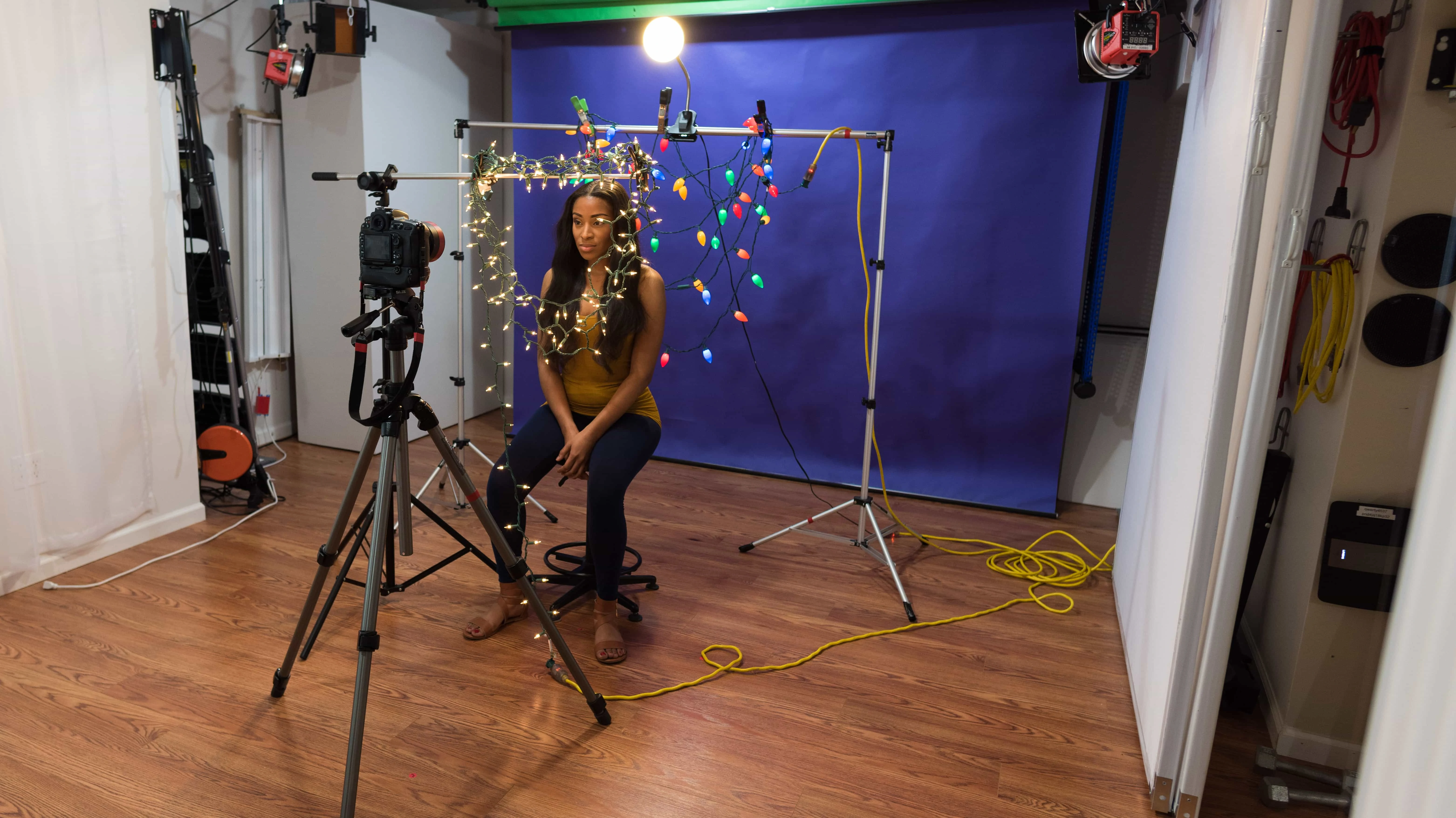 Behind the scenes shot of DIY Christmas light portraits model Monae with boom arm holding white Christmas lights in front of her and color bulbs behind her with desk lamp above her aimed down on same frame with purple background