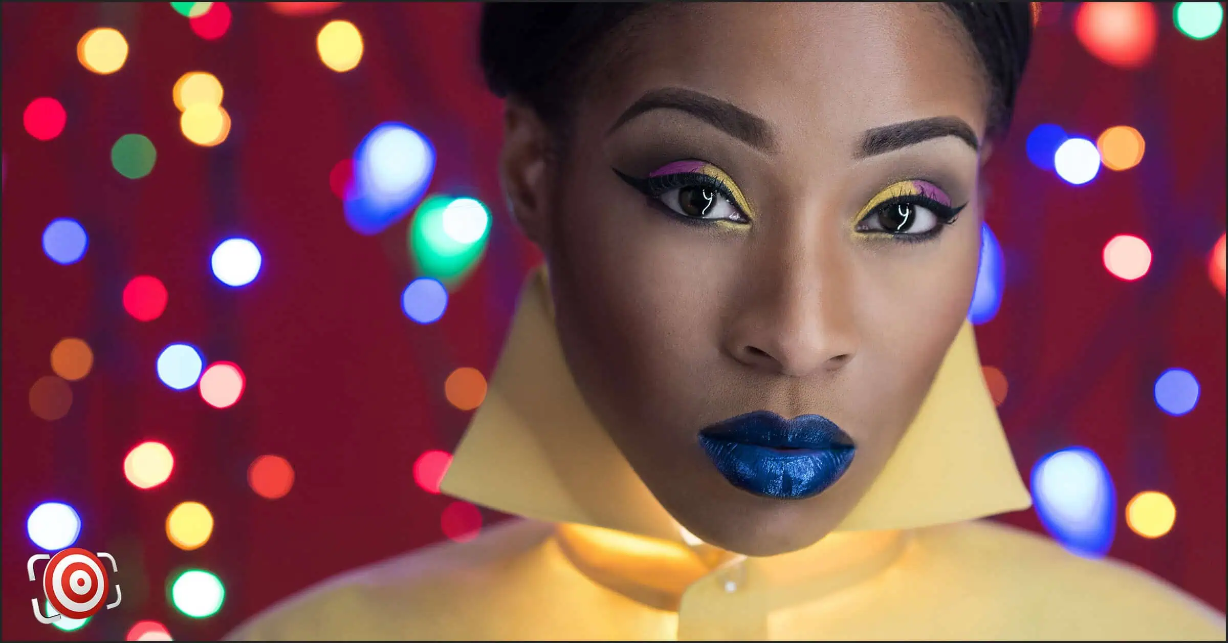 A woman with bold makeup, including blue lipstick and colorful eye shadow, wears a yellow collar against a background of colorful, out-of-focus lights, perfect for DIY Christmas light portraits.