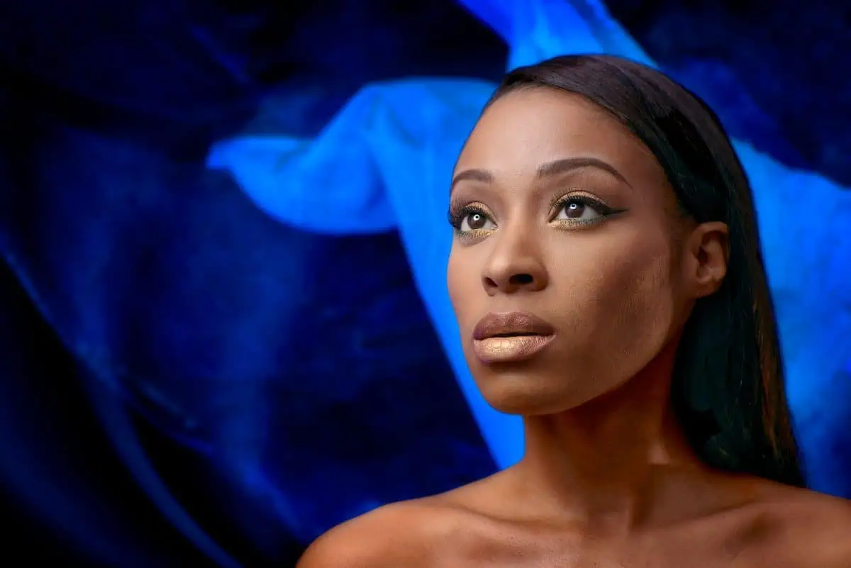 A woman with glowing skin and styled makeup gazes upward, beautifully illuminated by a beauty dish against a vivid blue fabric background.