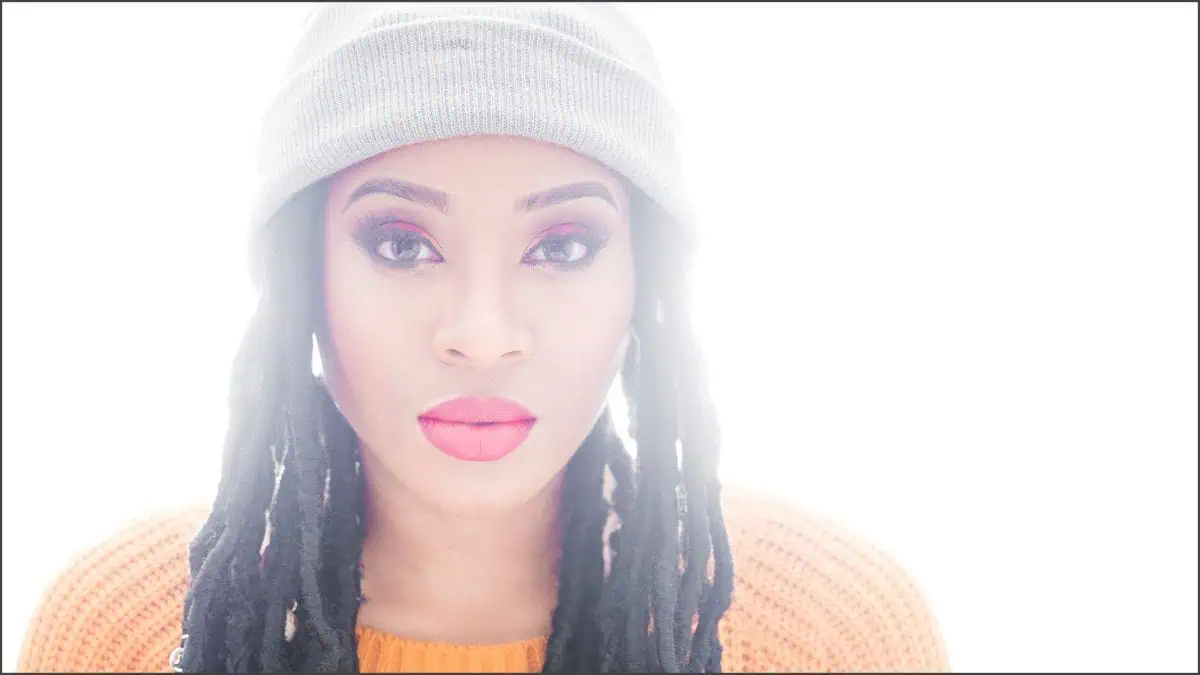 A person with long dark hair and a gray beanie, wearing a bright orange sweater and vivid makeup, poses against a white background. The setup captures their vibrant look as if lit perfectly by MagMod accessories.