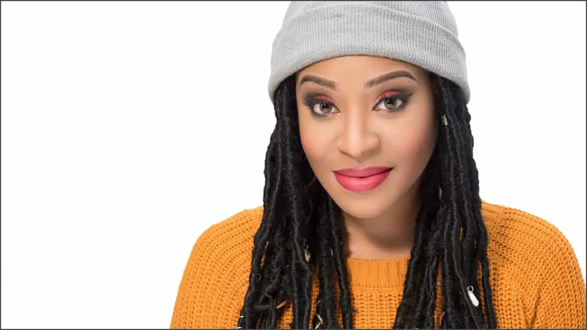 A person dons a grey beanie and an orange sweater, their long braided hair and red lipstick adding vibrancy. They smile slightly against a white background, embodying the perfect blend of style and charm reminiscent of a magmod photoshoot.