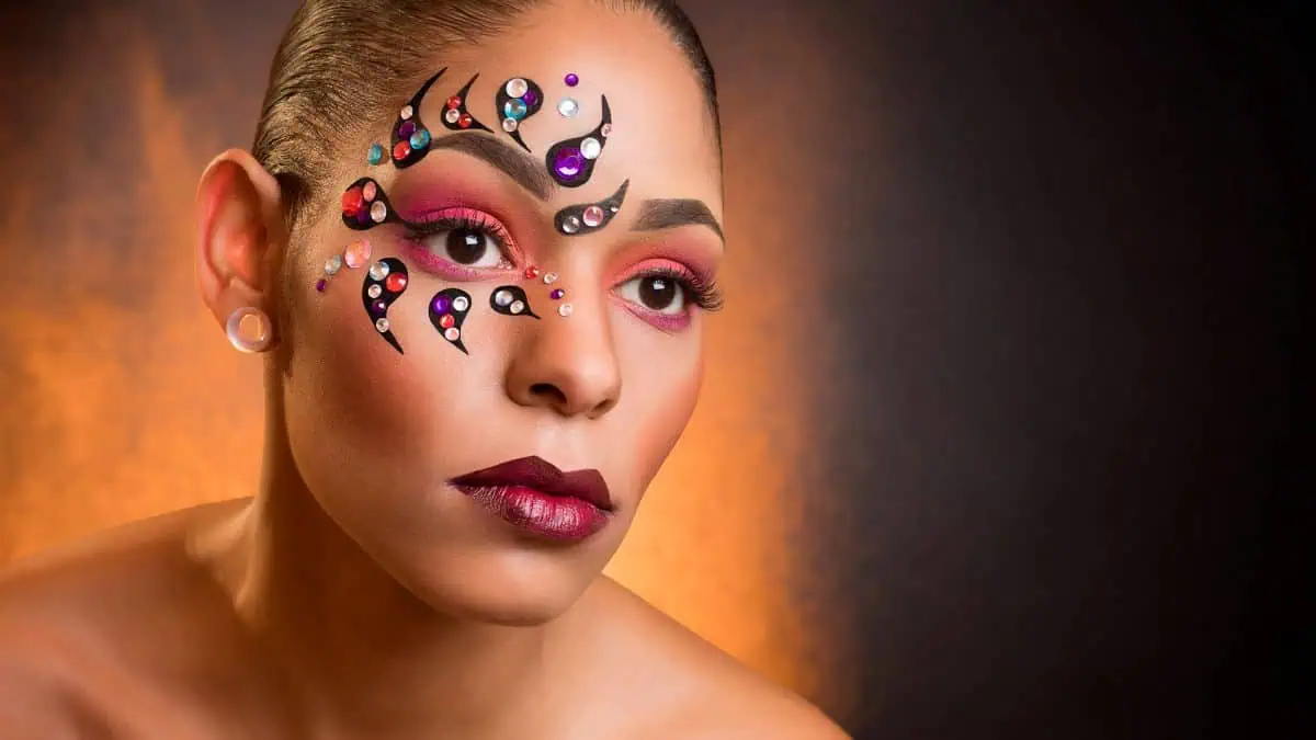 Person with artistic face paint adorned with colorful gemstones and black swirls, complemented by pink and burgundy makeup, poses against a gradient brown and orange background. Perfect for those exploring DIY photography tips to capture intricate details.