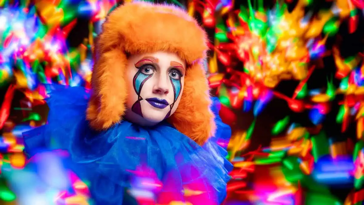 A person in colorful clown makeup and an orange hat, with a blue tulle collar, stands amidst bright, multicolored lights captured in a mesmerizing live composite.