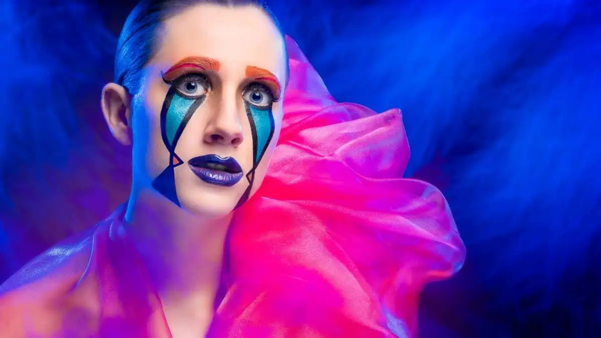 A person adorned with colorful, geometric makeup and striking blue lipstick stands enveloped in sheer, pink fabric. The scene unfolds against a mesmerizing blue smoky background, resembling a live composite of art and fashion brilliance.