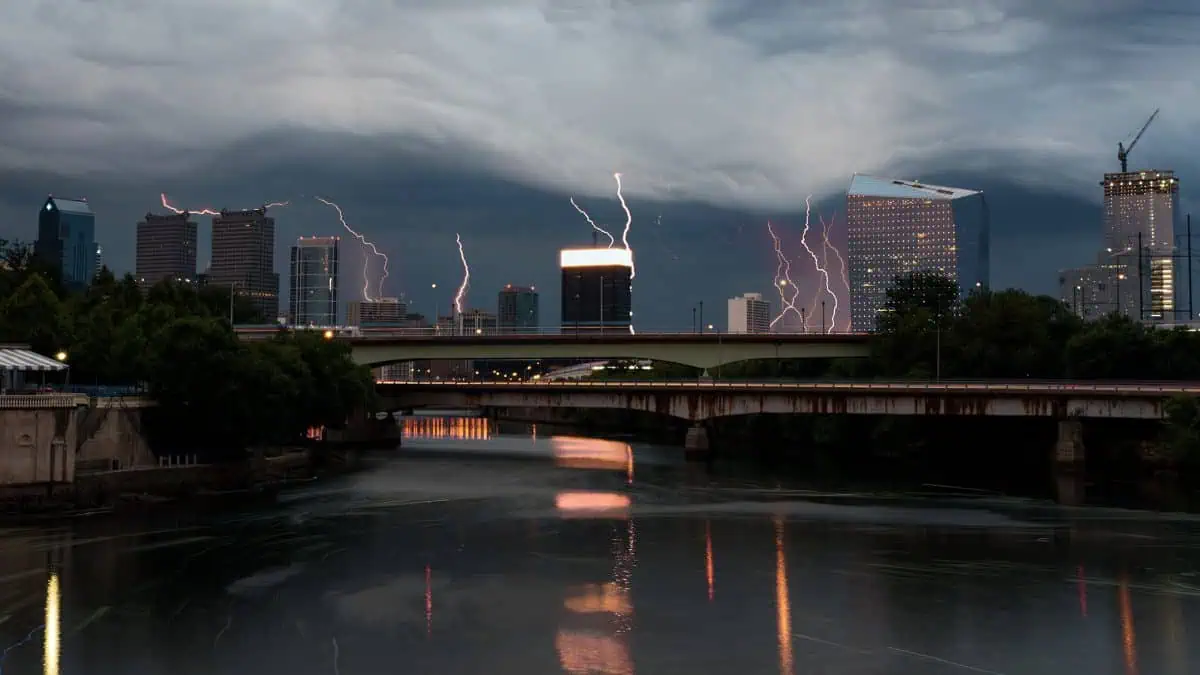A city skyline under a stormy sky with lightning bolts striking behind tall buildings, reflected in the calm river below, creates a mesmerizing live composite of nature's fierce beauty against urban serenity.