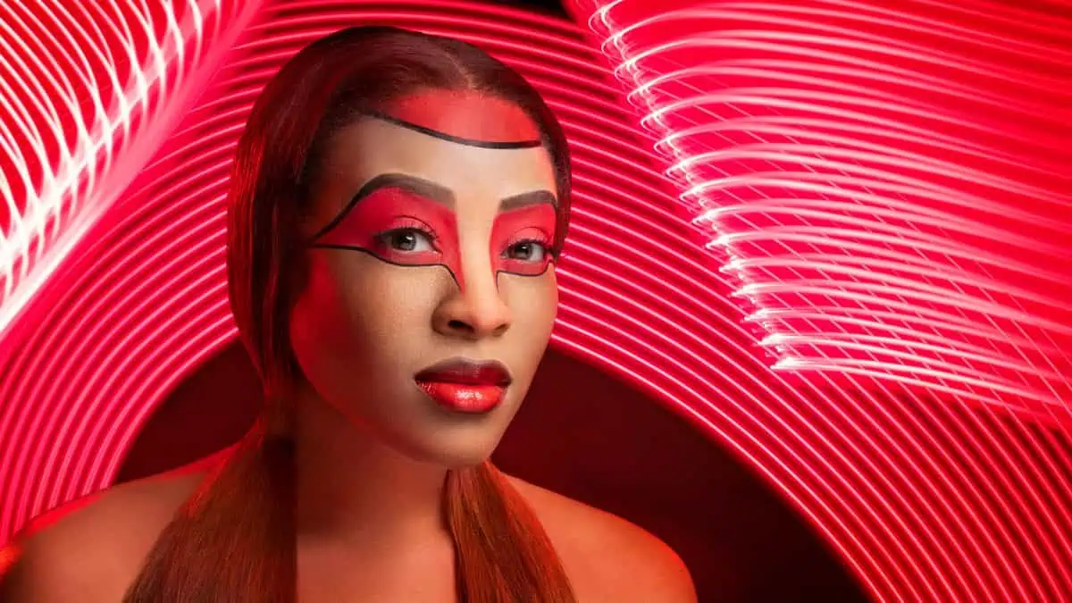 A person adorned with abstract red and black face paint emerges against a mesmerizing backdrop of swirling light patterns, creating an effect akin to a live composite masterpiece.