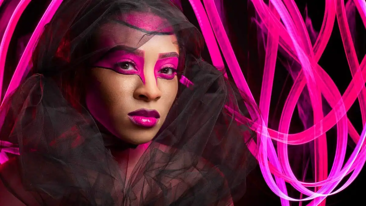 A person with vibrant pink makeup and a black veil stands elegantly, framed against a backdrop of swirling neon pink lights, creating a mesmerizing live composite.