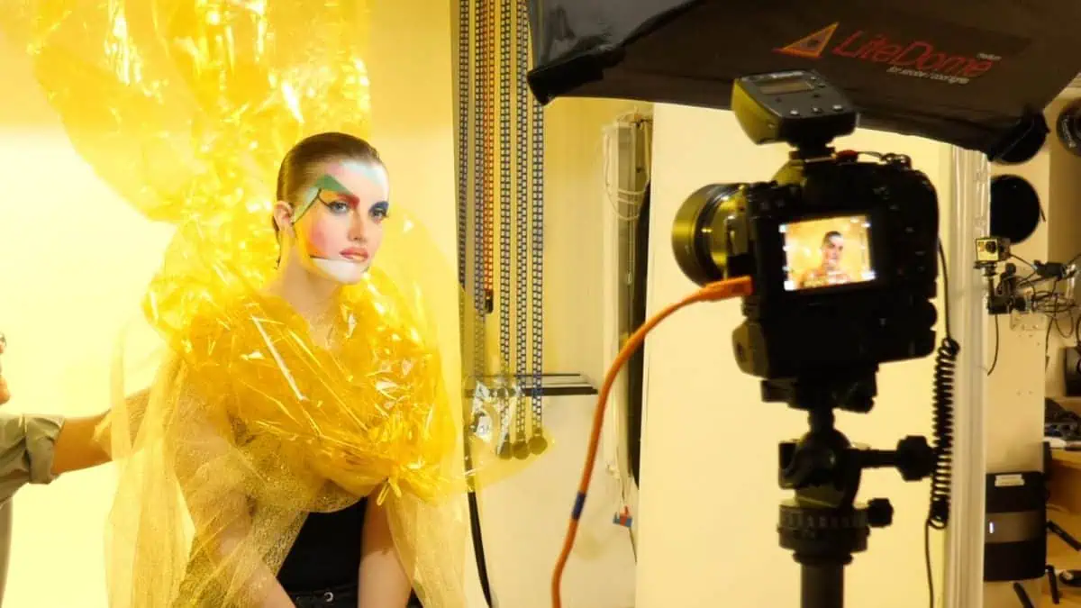 Model in an elaborate yellow wrap and colorful makeup poses for a photoshoot, seamlessly blending fashion portraits with cellophane. The vibrant scene features camera and lighting equipment, adding a dynamic touch to the creative atmosphere.