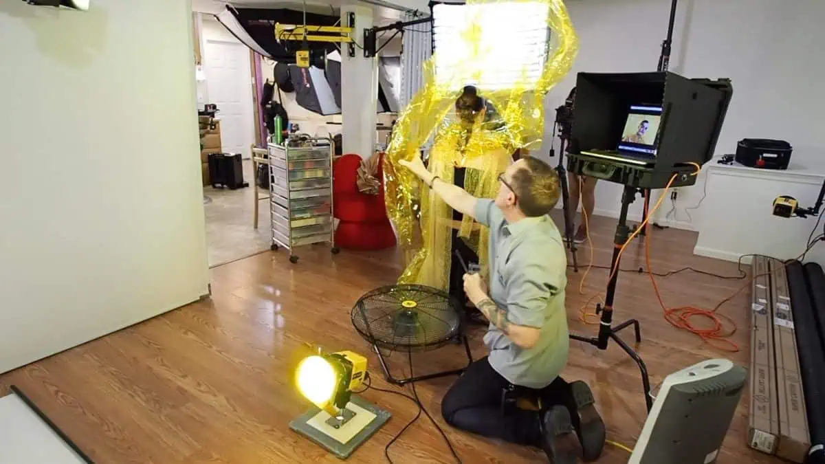 A photographer sets up a fashion portrait session with a model draped in shimmering gold plastic wrap. The studio lights, fan, and other equipment create a dynamic backdrop on the hardwood floor, enhancing the creative use of cellophane in this striking visual composition.