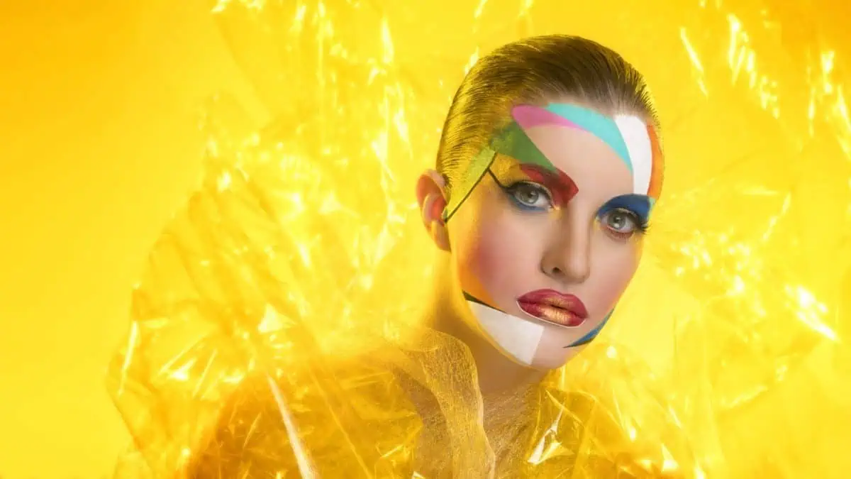 A striking fashion portrait features a person adorned with colorful geometric face paint, wrapped in translucent cellophane, set against a vibrant yellow background.