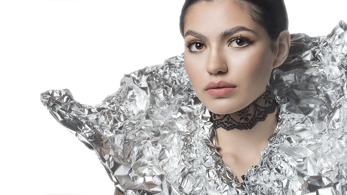 A woman with a neutral expression, highlighted by simple lighting, wears a black lace choker and a crinkled silver metallic garment against a white background.