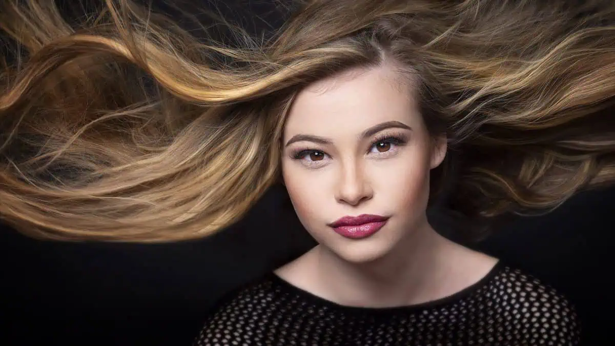 A woman with long, flowing hair captures the essence of blowing hair photography against a dark background. Her mesh top complements her serene expression as she gazes directly at the camera.