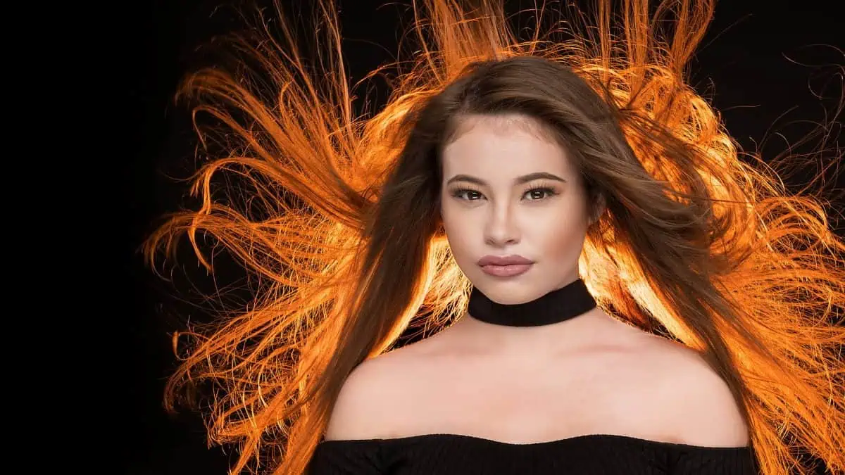 A woman with long hair stands against a black background, her hair blown back in an artistic display. It's a striking moment of blowing hair photography, with each strand catching the fiery glow behind her head.
