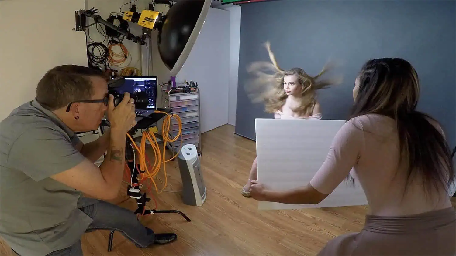 The photographer captures an image of a woman with blowing hair in a studio while an assistant holds a reflector.
