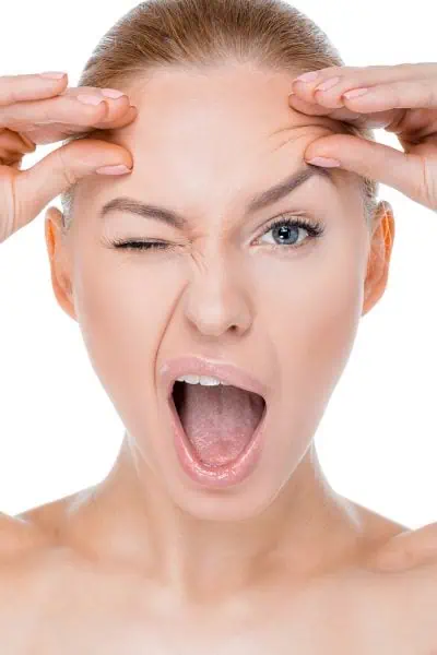 A person, seemingly caught blinking in photographs, holds their forehead with both hands, one eye closed and mouth open against a white background.
