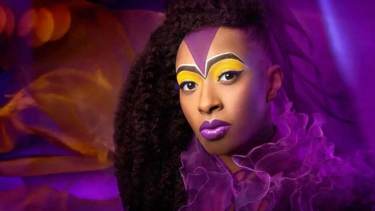 A woman with purple and yellow makeup is posing in front of a purple background.
