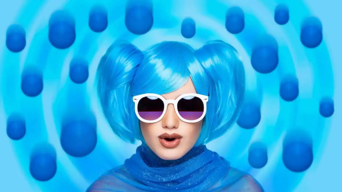 A woman with blue hair and sunglasses in front of a blue background.