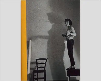 A black and white photograph of a woman standing next to a chair, taken by Bruno Barbey.