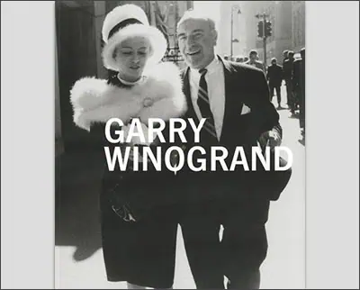 Black and white photo of a woman and a man walking on a city street, the man smiling and the woman dressed in a white fur and hat, with "garry winogrand" text overlay.