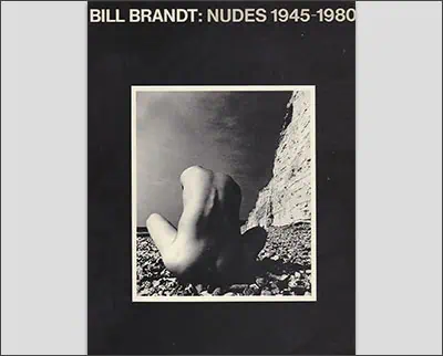 Cover of "Bill Brandt: Nudes 1945-1980" with a black-and-white photograph of a nude figure sitting on a rocky shore with a cliff in the background.