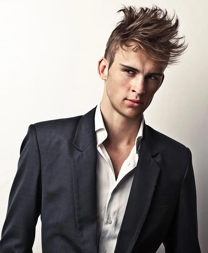 A man with tousled hair, embodying classic modeling terms, stands gracefully in a dark suit and white shirt against a plain background.