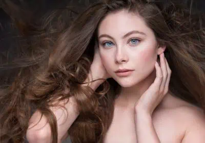 A person with long, flowing brown hair poses with hands near their face, gazing directly at the camera.
