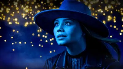 Woman in a wide-brimmed hat gazes upward in a blue-tinted image, with blurred golden lights in the background.