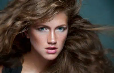 Close-up of a person with long, wavy brown hair and blue eyes, looking directly at the camera against a dark background.