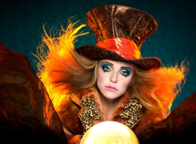 Person dressed in a colorful fantasy costume with a large top hat, vibrant makeup, and feathered details, holding a glowing orb.