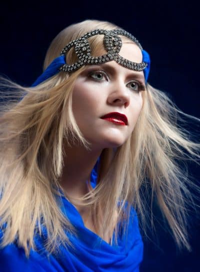 A woman with light hair wears a blue outfit and a decorative headband against a dark background.