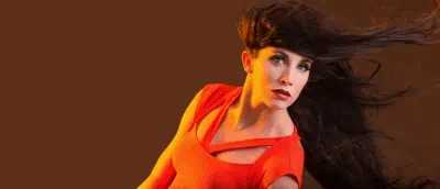 A woman with long flowing hair wears a bright orange top against a brown background.